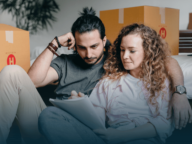 Et par som sitter i sofaen og ser på papir med flyttesker i bakgrunnen.